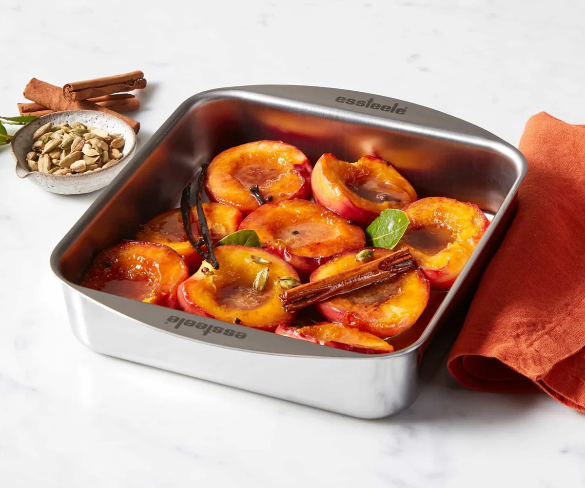 Essteele Clad Ovenware large rectangular stainless steel roasting pan, available at Chef&rsquo;s Hat, shown holding roasted stone fruit in syrup with cinnamon sticks, cardamom pods and bay leaves, styled with an linen napkin and spices on a marble surface.