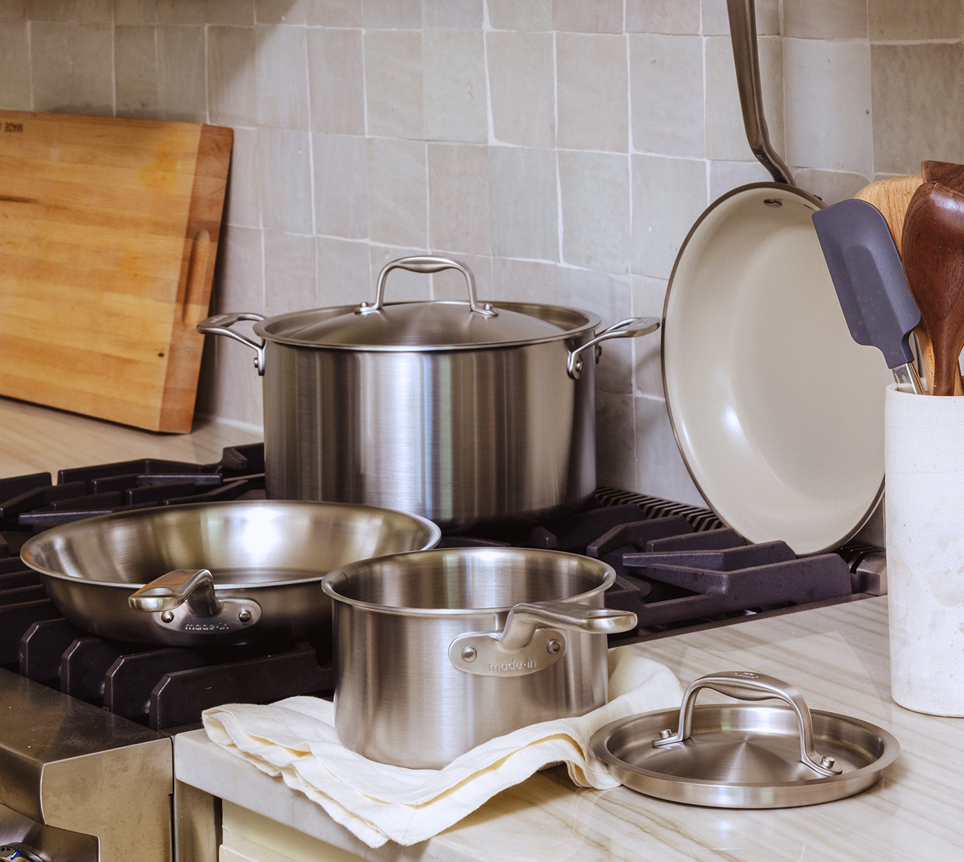 A set of Made In Stainless Clad and Ceramiclad Cookware is now available at Chef&rsquo;s Hat, arranged on a stovetop. A wooden cutting board and utensils are nearby, evoking a homey kitchen feel.