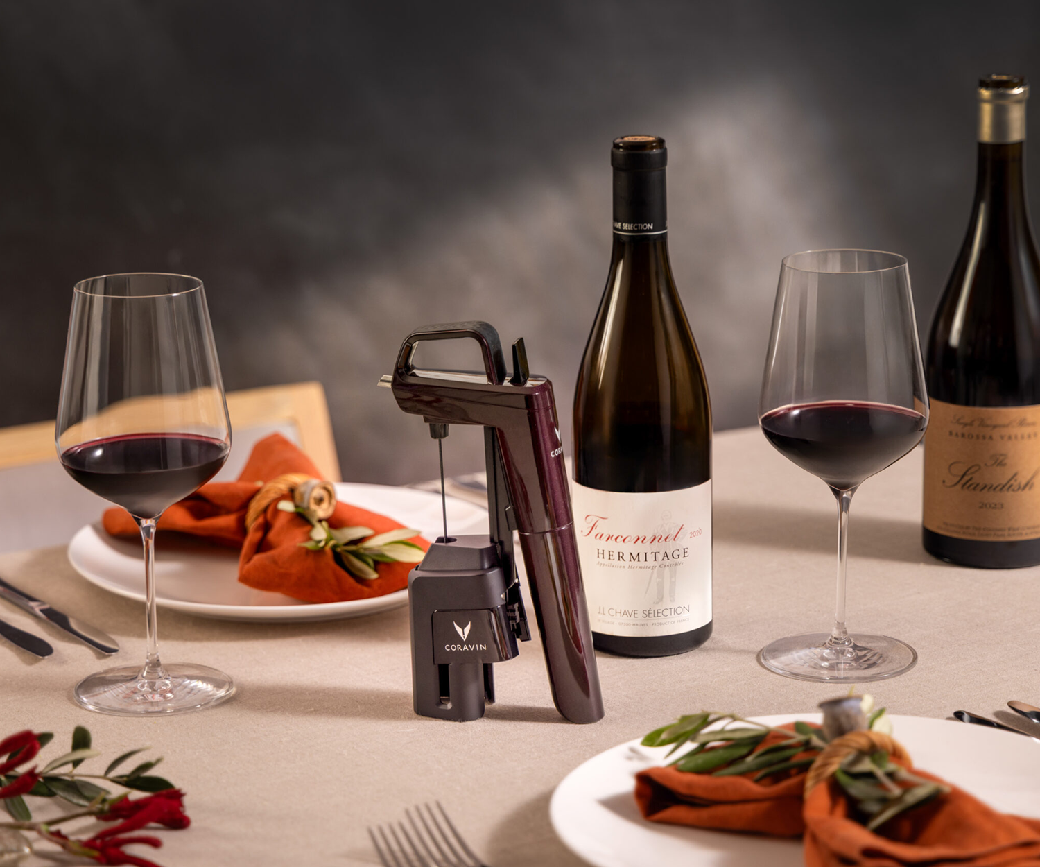 Tabletop featuring the Coravin Timeless Six+ wine by-the-glass system, beside a bottle of wine, a glass of red wine, and a styled background with plates of food for a refined dining setting.