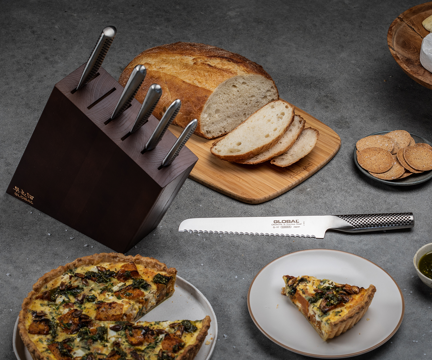 Bread and cheese on a grey surface beside a Global knife block set made from CROMOVA 18 stainless steel. A sliced loaf sits on a wooden board, with cheese on a stand and olives nearby, creating a rustic dining feel.