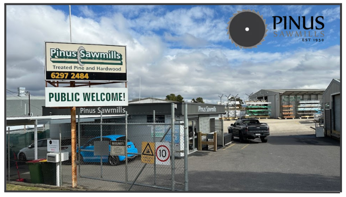 Entrance of Pinus Sawmills, showing the main gate, signage welcoming the public, and visible storage areas for treated pine and hardwood products.